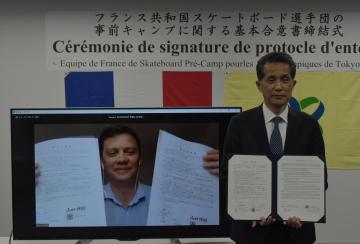 東京五輪の事前キャンプに関する基本合意書に署名した山口伸樹市長とフランスのローラー・スケートボード連盟のボリス・ダーレット会長(画面)=笠間市役所