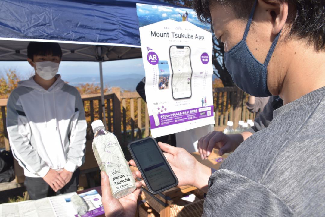 筑波山登山道アプリを示して記念品の飲料水を受け取った実証実験の利用者=つくば市筑波の筑波山
