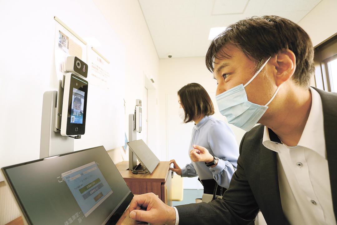 社屋入り口に設置された顔認証の体温測定器に顔を近づける東京フードの社員＝つくば市上大島
