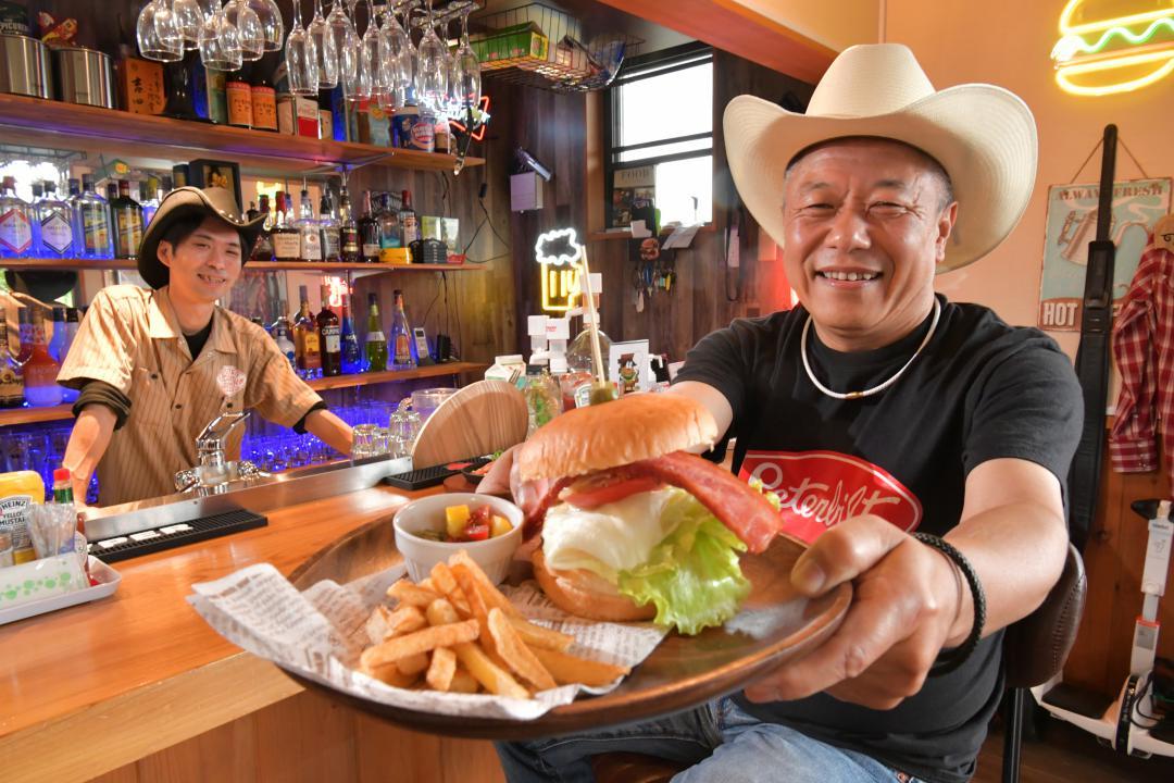 アメリカ南部のバーの雰囲気が漂う店内。自慢のナッシュビルハンバーガーを持つ京免義典さん(右)=常陸太田市大里町
