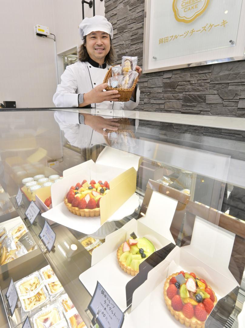 地元特産の果物をふんだんに使ったチーズケーキが並ぶショーケース。代表の茂垣圭輝さんが商品を手に笑顔を見せる=鉾田市田崎
