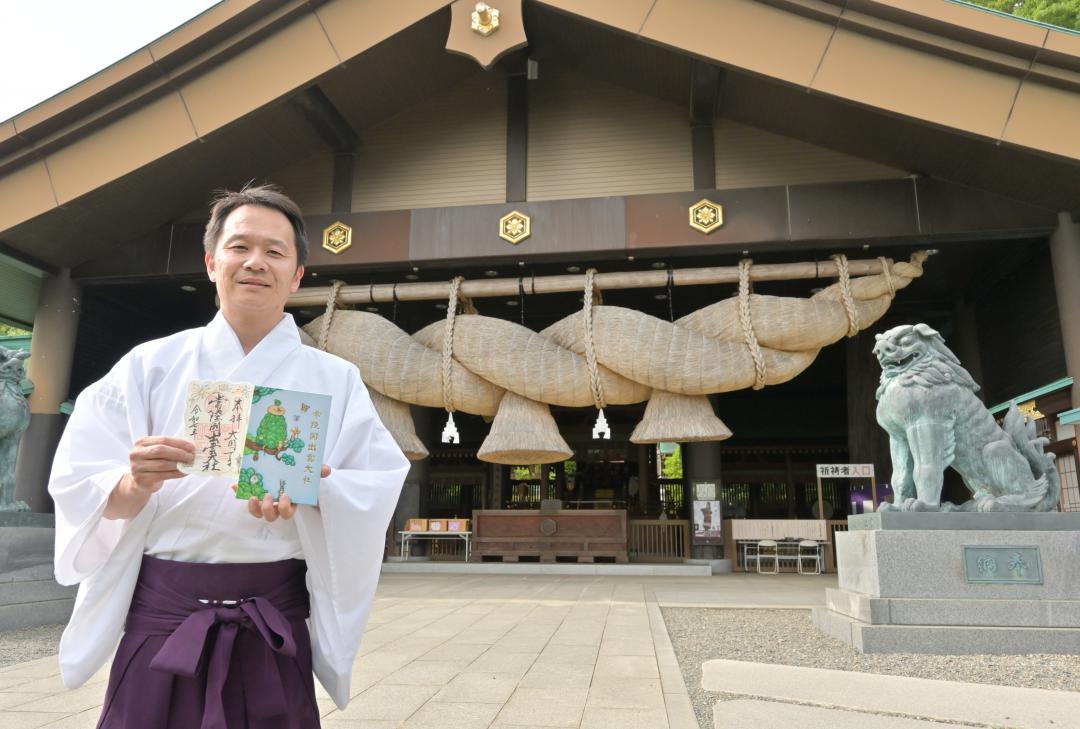 巨大なしめ縄をつるした拝殿の前で、限定御朱印と御朱印帳を持つ高橋正重権宮司=笠間市福原
