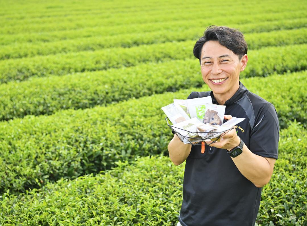 夏の芽が出始めた茶畑で、さしま茶を活用したパウンドケーキなどを手に笑顔を見せる松田製茶の松田浩一代表=八千代町水口
