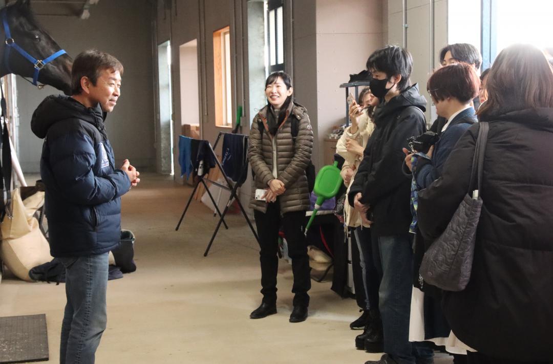 厩舎で手塚貴久調教師(左)から説明を受けるツアー参加者=美浦村美駒